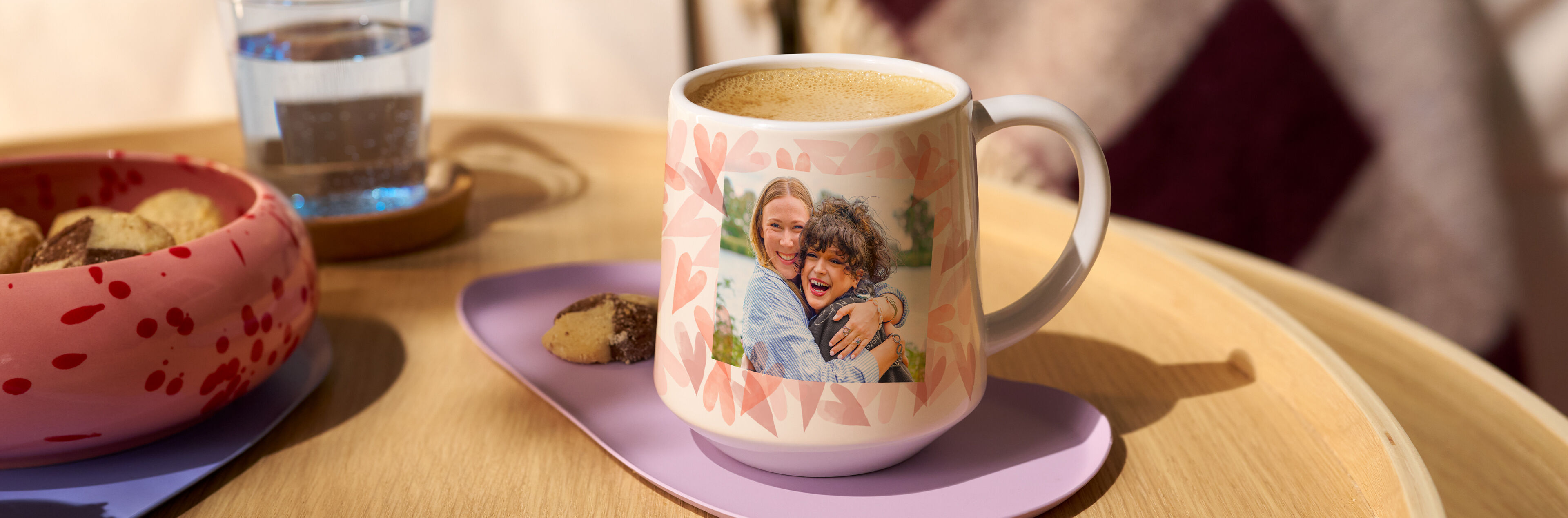 Personalisierte Tasse mit Foto von zwei lachenden Freundinnen auf einem Tisch mit Kaffee und Gebäck.