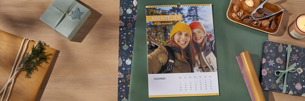 Ein Fotokalender liegt auf einem Tisch mit Geschenkpapierbögen, einem verpackten Geschenk, einer Kerze und einer Schale mit Nüssen. Das Kalenderblatt Dezember zeigt das Foto von zwei lachenden Frauen in winterlicher Kleidung sowie die Worte “Abenteuer willkommen”.