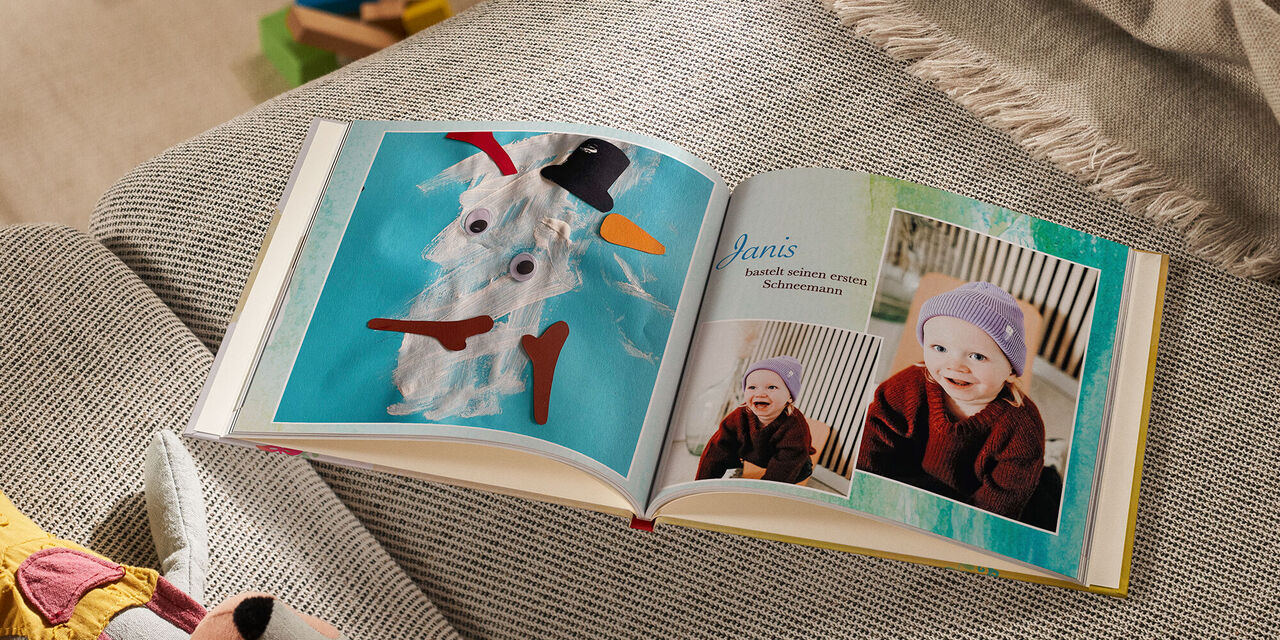 Auf einem Sofa liegt ein geöffnetes CEWE FOTOBUCH neben einem Kuscheltier und einer Decke. Die linke Seite zeigt das Foto einer Kinderbastelarbeit, die einen Schneemann darstellt. Auf der rechten Seite sind zwei Fotos von einem lachenden Kind zu sehen. Die Überschrift lautet „Janis bastelt seinen ersten Schneemann“. Der Hintergrund der Doppelseite ist mit blau-grünen Aquarellfarben ausgefüllt.