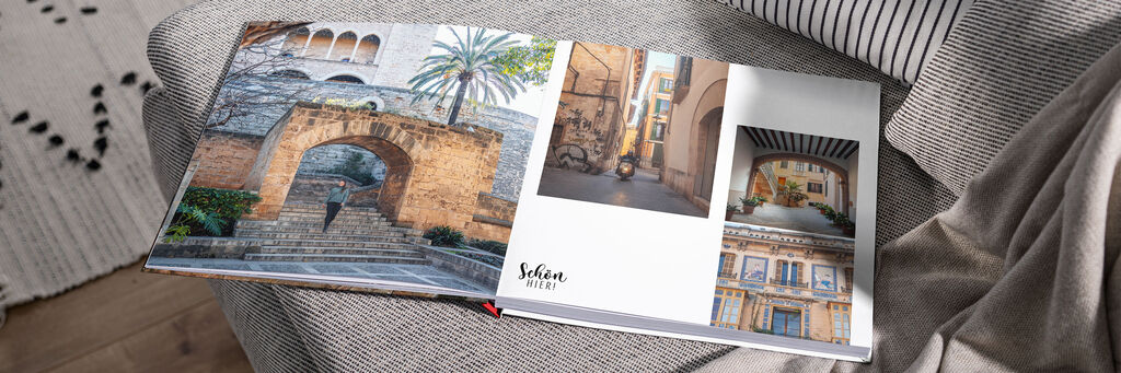 Auf der Doppelseite eines Fotobuchs sind vier Bilder von einer Altstadtkulisse auf Mallorca zu sehen. Auf der linken Seite sieht man eine Frau eine Treppe hinabsteigen, im Hintergrund Palmen. Das CEWE FOTOBUCH liegt aufgeschlagen auf einem Sofa in Wohnumgebung.