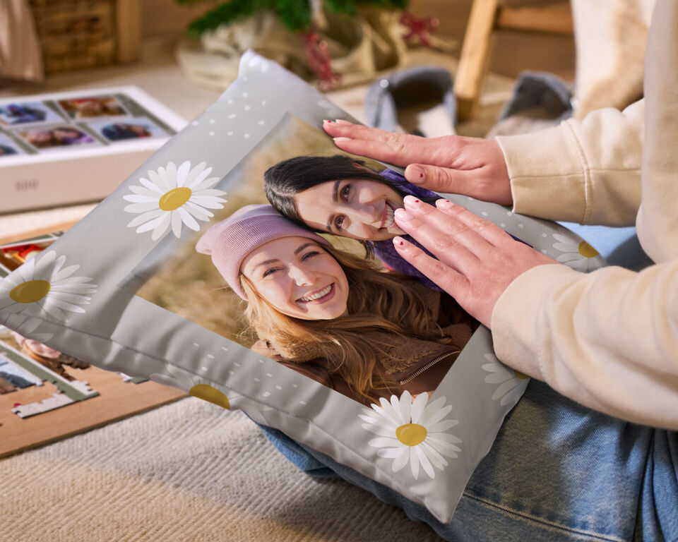 Zwei Hände halten ein graues, bedrucktes Kissen mit Blumenmuster und dem Foto zweier lachender Frauen, im Hintergrund ein teilweise zusammen