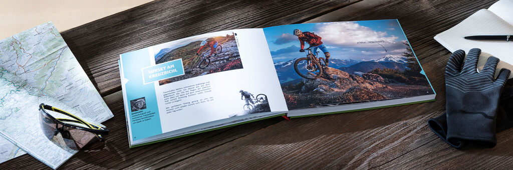 Auf einem Tisch liegen ein Fotobuch, Handschuhe, ein Heft und Landkarten. Auf dem Fotobuch sind ein Mountainbike sowie die Aufschrift “Alpentrail 2018” zu sehen.