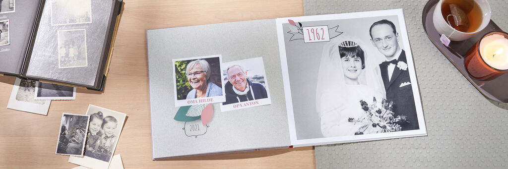 Auf einem Tisch liegt ein aufgeschlagenes CEWE FOTOBUCH. Darin sind Fotos von Großeltern zu sehen. Links sind aktuelle Fotos, rechts ein Foto vom Hochzeitstag 1962. Auf dem Tisch liegen alte Schwarzweißfotos und neben dem Fotobuch steht ein Tablet mit Kerze und Tee.