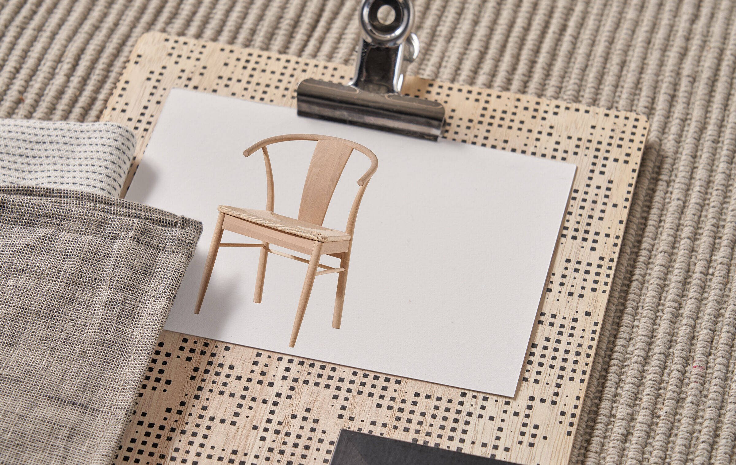 Auf einem Stück Stoff aus geriffelt gewebter Naturfaser liegt ein Klemmbrett aus Holz mit kleinem schwarz-cremefarbenem Quadratmuster. Auf das Brett ist ein Foto geklemmt, das einen beigefarbenen Stuhl zeigt.